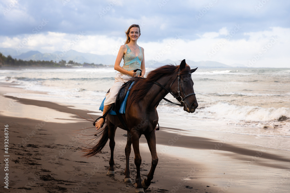 Beautiful woman riding horse on the beach. Outdoor activities. Caucasian woman wearing skirt. Traveling concept. Cloudy sky. Sea view. Copy space. Horizontal layout. Bali, Indonesia Beautiful woman riding horse on the beach. Outdoor activities. Caucasian woman wearing skirt. Traveling concept. Cloudy sky. Sea view. Copy space. Horizontal layout. Bali, Indonesia