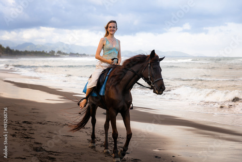 Beautiful woman riding horse on the beach. Outdoor activities. Caucasian woman wearing skirt. Traveling concept. Cloudy sky. Sea view. Copy space. Horizontal layout. Bali, Indonesia