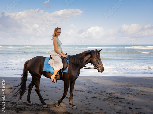 Beautiful woman riding horse on the beach. Outdoor activities. Caucasian woman wearing skirt. Traveling concept. Cloudy sky. Sea view. Copy space. Horizontal layout. Bali  Indonesia