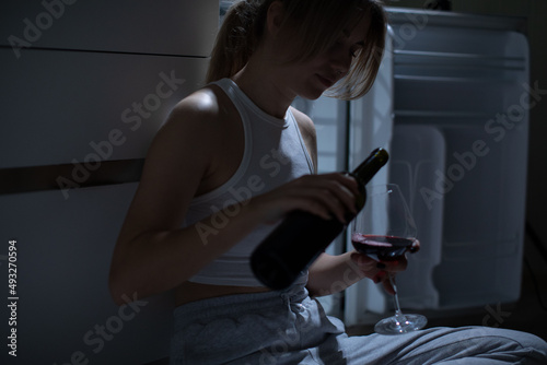 Sad woman drinking wine sitting in front of an open refrigerator in the dark.