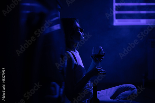 Sad woman drinking wine while sitting near the refrigerator in the dark.