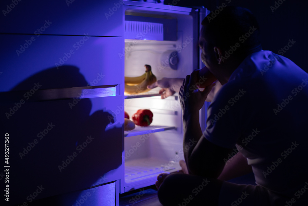 A man with a sausage in his mouth sits by an open refrigerator in the middle of the night. A man with a sausage in his mouth sits by an open refrigerator in the middle of the night.