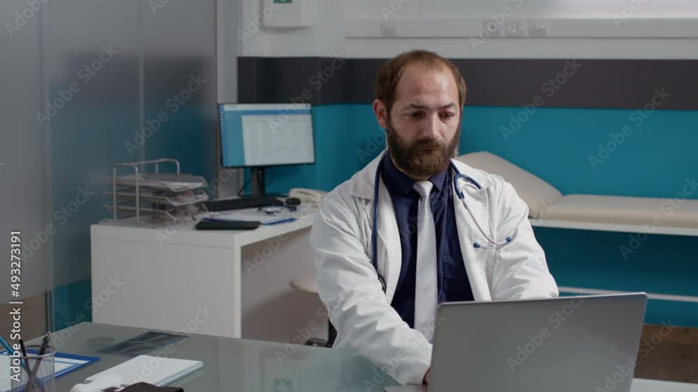 Portrait of general practitioner working on laptop in medical office, wearing white coat. Male physician using computer to help patients with disease, having healthcare expertise. Portrait of general practitioner working on laptop in medical office, wearing white coat. Male physician using computer to help patients with disease, having healthcare expertise.