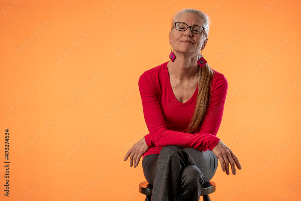 Beautiful smiling happy blond mature woman wearing eyeglasses on solid orange background with copy space sitting on stool. Beautiful smiling happy blond mature woman wearing eyeglasses on solid orange background with copy space sitting on stool.