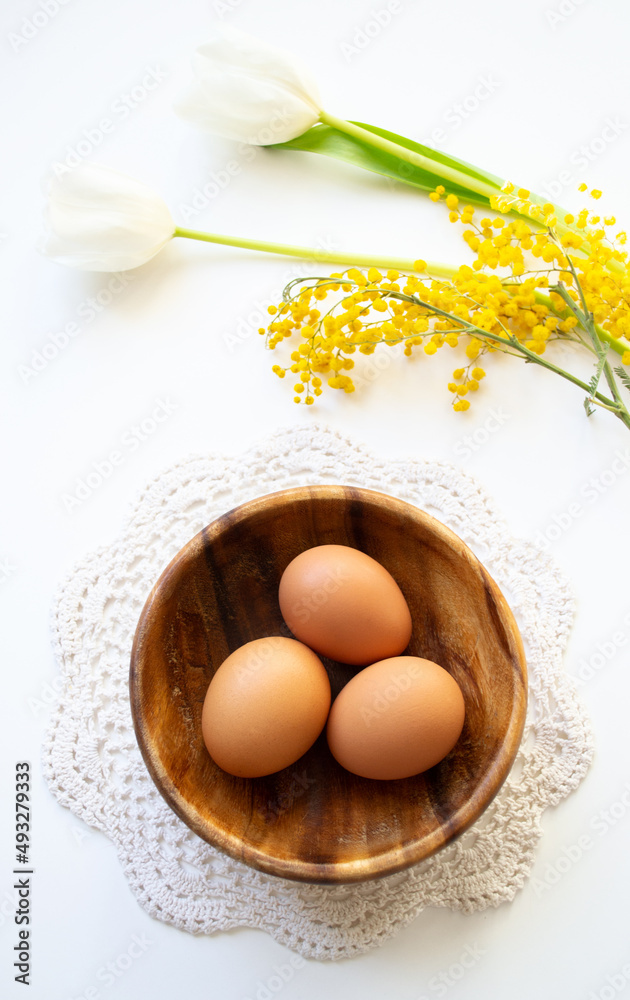 Brown raw chicken eggs in a wooden plate on a lace doily. Easter banner with copy space with tulips and mimosa on white background