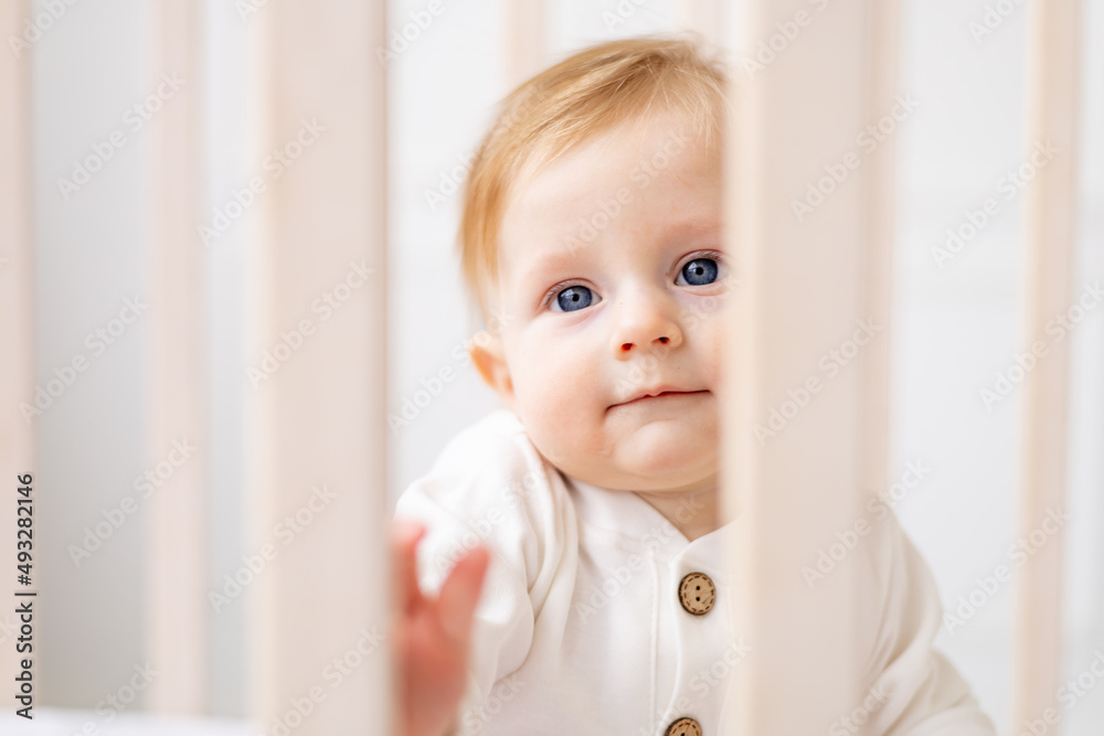 close-up portrait of a baby 6 months old, a blond boy with blue eyes in a crib in a bright bedroom in a white cotton bodysuit, the concept of children's goods