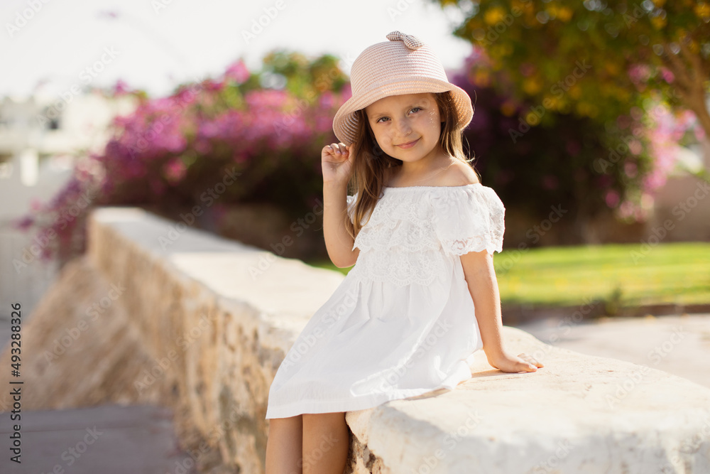 Portrait of a little happy girl with blue eyes in a straw hat and white dress with hat shadows on her face sitting on the background of a tropical resort. Concept of childhood and summer vacation.