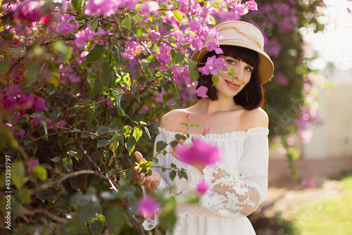 Portrait of a charming young woman in a white dress and straw hat standing near a blooming pink tree on a sunny spring day. Beautiful caucasian young woman at flowered garden. © sofiko14