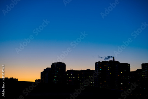 The dark outlines of city buildings against a clear evening dark blue sky. Evening yellow-blue sky gradient. Evening in city. Night city. City landscape background.
