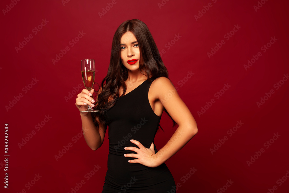 Beautiful brunette model stands on a red background and smiles a beautiful smile in a white dress, she holds a glass of champagne Beautiful brunette model stands on a red background and smiles a beautiful smile in a white dress, she holds a glass of champagne