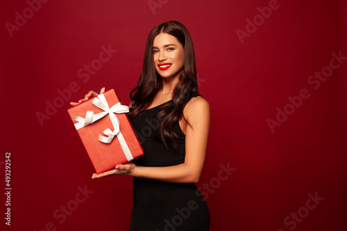 beautiful brunette model stands on a red background and smiles a beautiful smile in little black dress, she holds a gift,