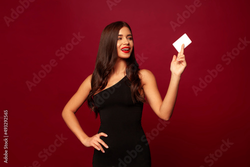 Beautiful brunette stands on a red background in a little black dress, with red lipstick and smiles, she holds a white credit card. Promotional photo for the theme of shopping, discounts, purchases