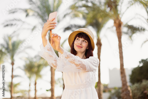 A woman in srtaw hat and white dress using smart phone or taking a selfie on the background with palm trees in summer. Holidays Vacation. Mobile Communication, Connection Concept.