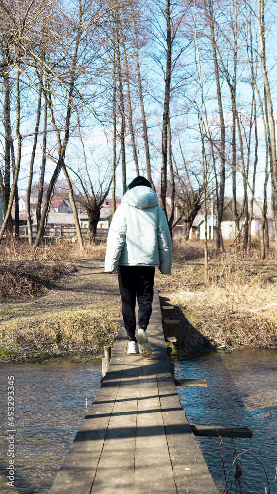 A girl in a blue jacket with a hood crosses a river in the forest on a makeshift bridge photo from the back