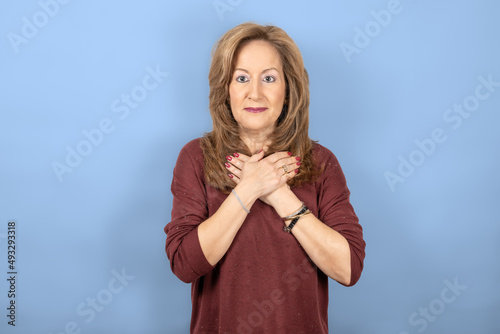 Woman with hands on chest excited in surprise, blue studio background.