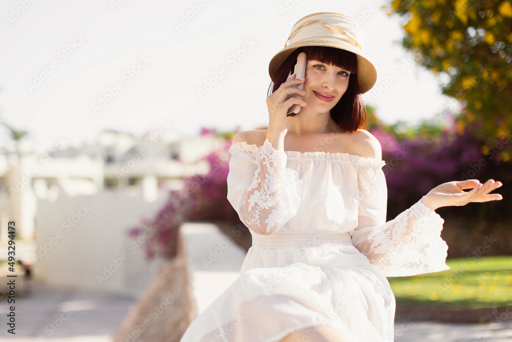 Happy cheerful woman talking on the phone during vacation. Smiling girl making answering call by cellphone sitting on the tropical background while having pleasant conversation speaking by mobile