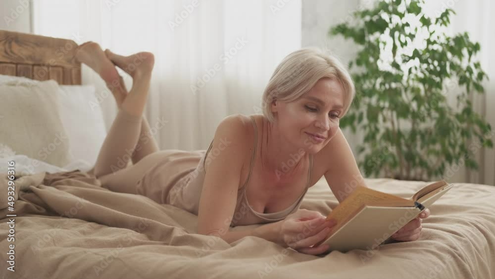 Interesting novel. Reading woman. Enjoying rest. Smiling middle-aged lady in sleeping suit laying with book on bed in light home interior. Interesting novel. Reading woman. Enjoying rest. Smiling middle-aged lady in sleeping suit laying with book on bed in light home interior.