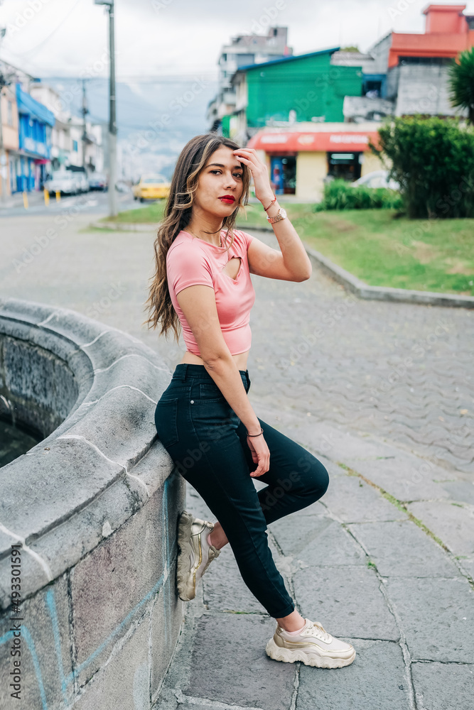 full body of attractive young latin woman standing leaning against stone wall, wearing makeup, casual clothes, long blonde hair, city lifestyle