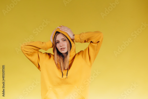 Portrait of sad young teen girl holding hands on head, wearing casual yellow hoodie in tone with background