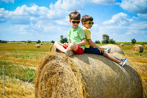 Two little boy stand among round haystack. Field with round bales after harvest under blue sky. Big round bales of straw, sheaves, haystacks