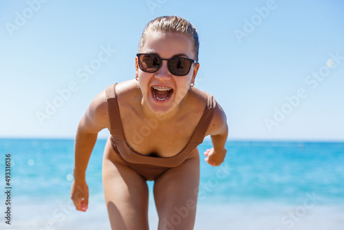 Tanned woman in sunglasses and a beige swimsuit having fun on the seashore and looking at the camera