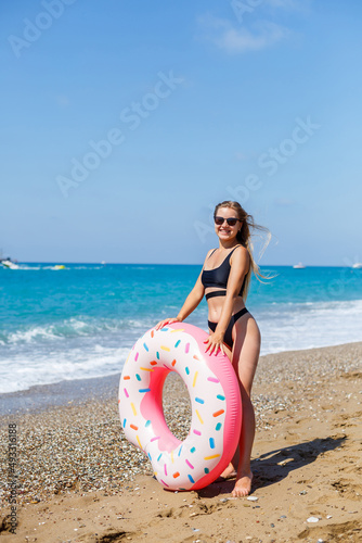 A slender woman in a stylish black swimsuit and sunglasses holds an inflatable circle donut in her hand. Beautiful happy girl posing and having fun in the sun. Vacation. travel by sea