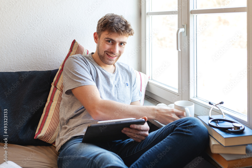 Junger Medizin-Student sitzt zu Hause mit einem digitalen Tablet auf seinem Bett vor einem Fenster und lernt. Er greift gerade zu eine Tasse und lacht in die Kamera. 