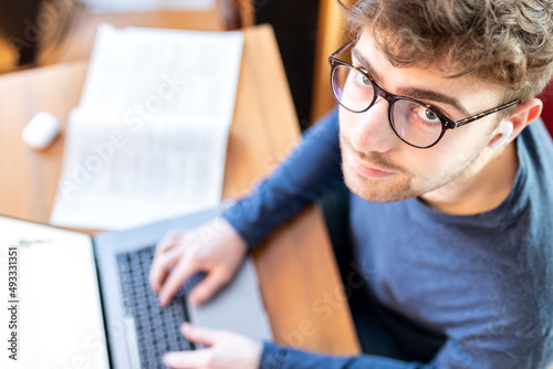 Junger Mann sitzt am Schreibtisch mit seinem Laptop und arbeitet, Blick von oben, er schaut in die Kamera.