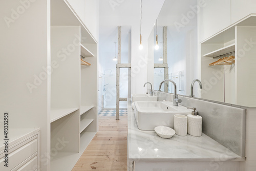 Bathroom of a bedroom with a dressing room with a white marble top with gray veining  a double white porcelain sink  a large long mirror and a shower in the background