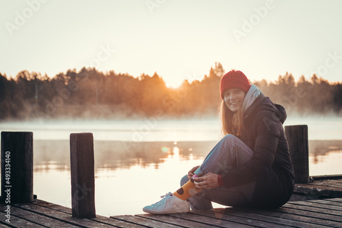 Photography Young brunette with a beautiful, realistic smile sits on a wooden bridge by a lake at sunrise