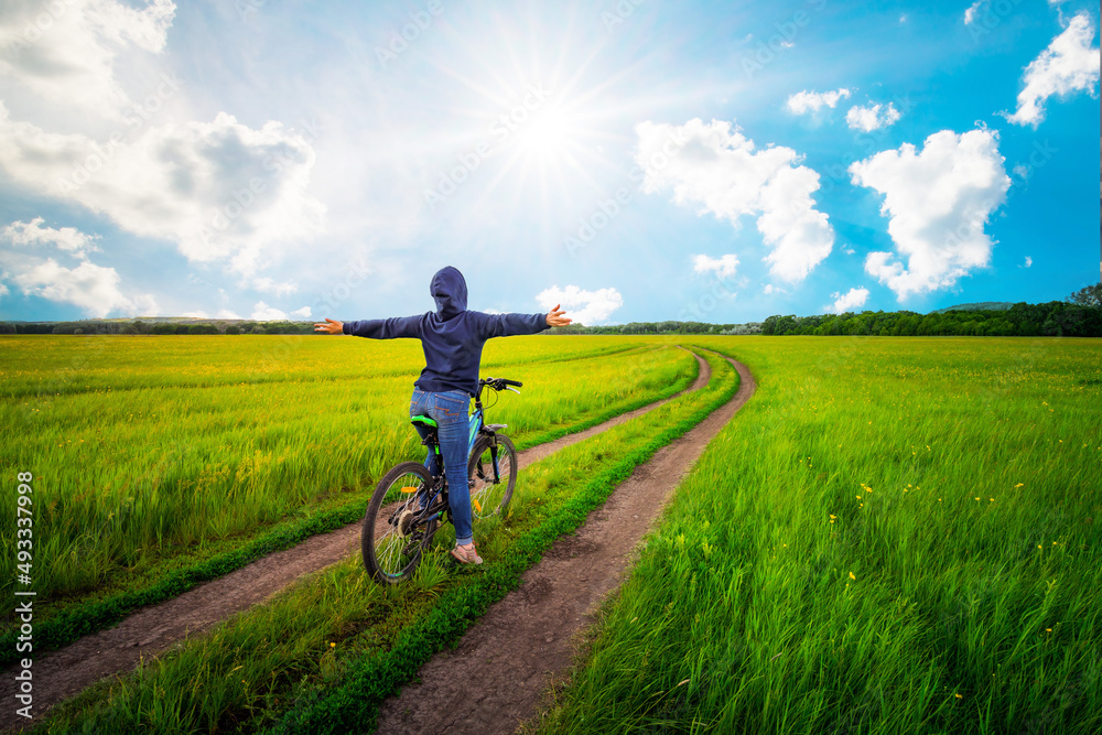 Woman with hoodie on a bike opens her arms by the gravel road. Sunny summer day and green grasses. Woman with hoodie on a bike opens her arms by the gravel road. Sunny summer day and green grasses.