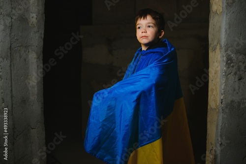 A sad Ukrainian boy wrapped in a Ukrainian flag, hiding from bombings in a bomb shelter in the city of Mariupol or Kiev. Russia's aggression. The war in Ukraine and 2022. © Serhii