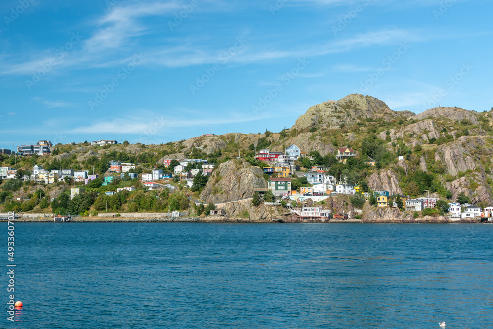 The hillside of St. John's Harbour, on a sunny day, under blue sky and white clouds. The colorful wooden houses are scattered along the hillside with the blue ocean in the foreground. The hillside of St. John's Harbour, on a sunny day, under blue sky and white clouds. The colorful wooden houses are scattered along the hillside with the blue ocean in the foreground.