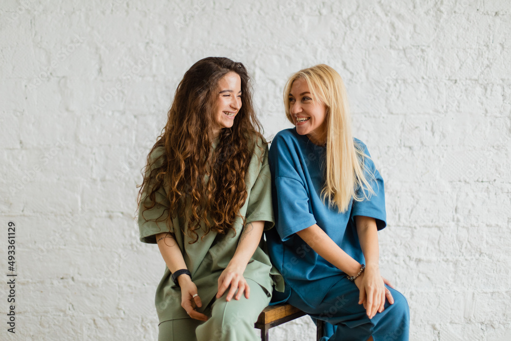 Two girls sit near white wall hugging each other Two girls sit near white wall hugging each other