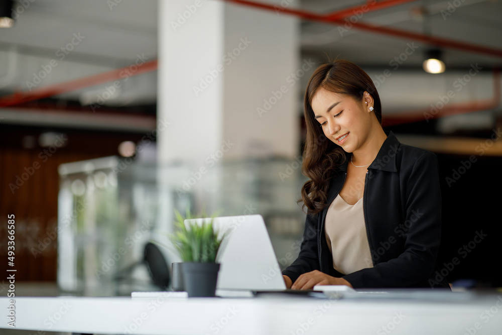 Smiling young Asian businesswoman working on a laptop computer at her desk in a bright modern home office, doing calculating expense financial report finance making notes on paper graph data document. Smiling young Asian businesswoman working on a laptop computer at her desk in a bright modern home office, doing calculating expense financial report finance making notes on paper graph data document.
