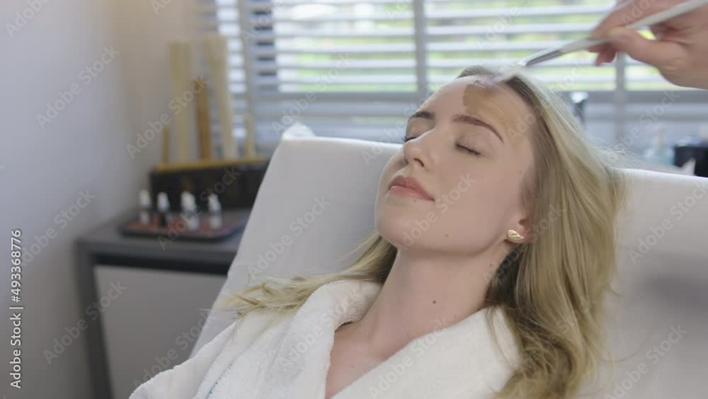 Young blonde caucasian woman receive a health skin care facial treatment while lying on a massage bed in luxury resort spa saloon. Skilled therapist hands apply with brush natural cream oils Young blonde caucasian woman receive a health skin care facial treatment while lying on a massage bed in luxury resort spa saloon. Skilled therapist hands apply with brush natural cream oils