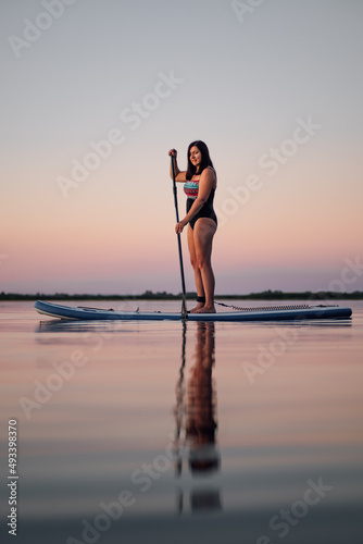 Middle age lady standing on blue sup board neat reeds holding oar in hands on lake looking away with pretty pink sky in background in evening. Active lifestyle for older people. © Юля Бурмистрова
