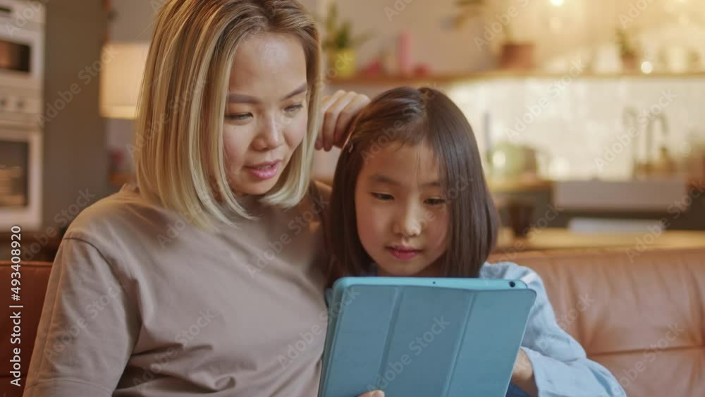 Happy Young Asian Mom and Daughter Doing Homework by Using Tablet, Device Draw With Pencel, Sitting on Sofa at Home. Young Korean Family Using Tad Together Happy Young Asian Mom and Daughter Doing Homework by Using Tablet, Device Draw With Pencel, Sitting on Sofa at Home. Young Korean Family Using Tad Together