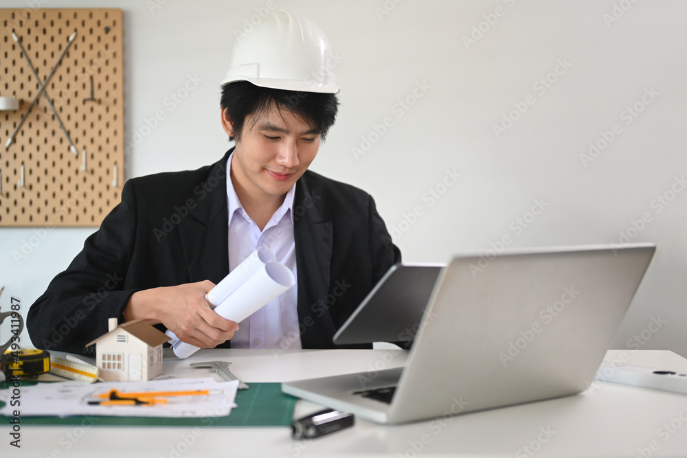 Handsome Architect man holding roll of architectural projects and working wit laptop in office.