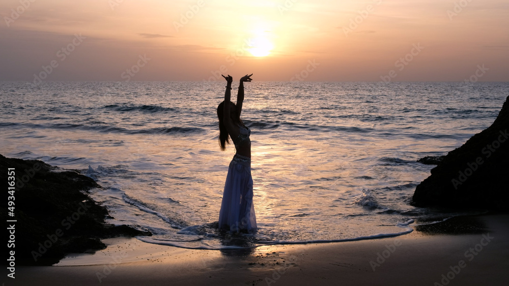 Woman dancing on the shore of the beach. Exotic woman looking at sunset at the horizon dancing belly dance on the beach. Woman raising hands dancing belly dance on the beach with the sunset. 
