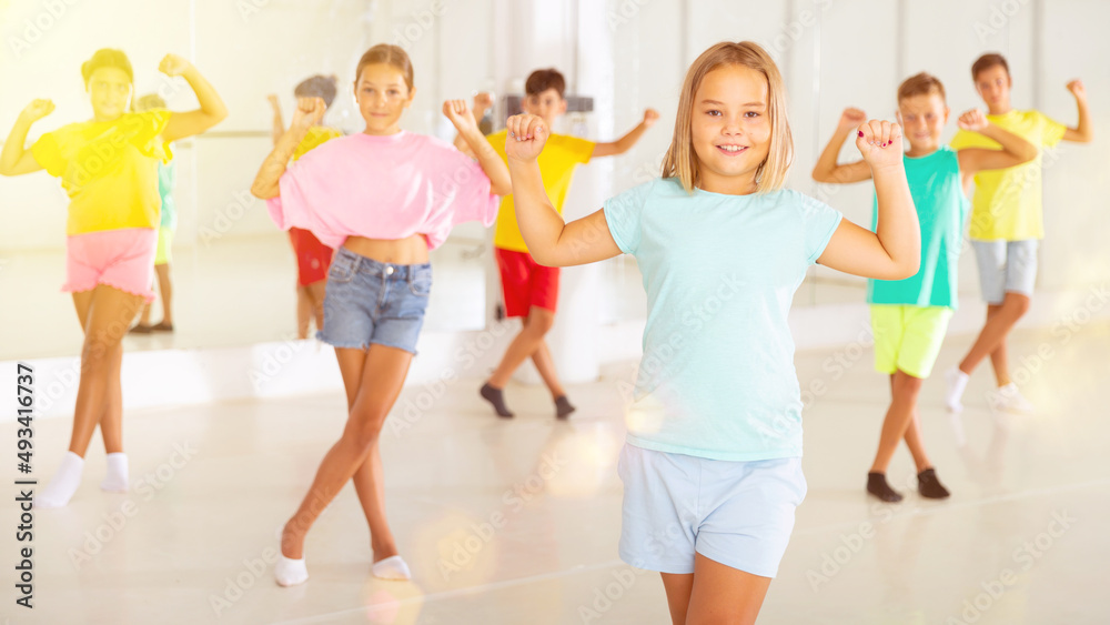 Smiling little girl training movements of modern vigorous dance with group of tweens in children dance studio.