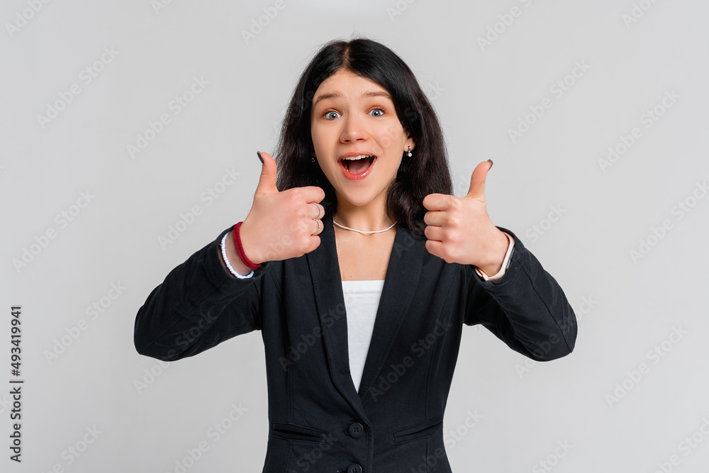 Cheerful happy pretty brunette teen girl showing thumbs up, smile recommend, wear in black blazer, standing over grey background. Young emotional woman Cheerful happy pretty brunette teen girl showing thumbs up, smile recommend, wear in black blazer, standing over grey background. Young emotional woman
