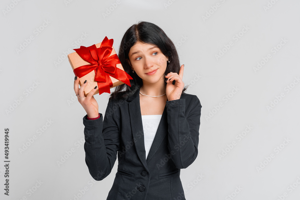 Thoughtful brunette teenager girl holding gift box, standing over white background intending to realizes the solution while lifting a finger up. Holidays, celebration and birthday concept Thoughtful brunette teenager girl holding gift box, standing over white background intending to realizes the solution while lifting a finger up. Holidays, celebration and birthday concept