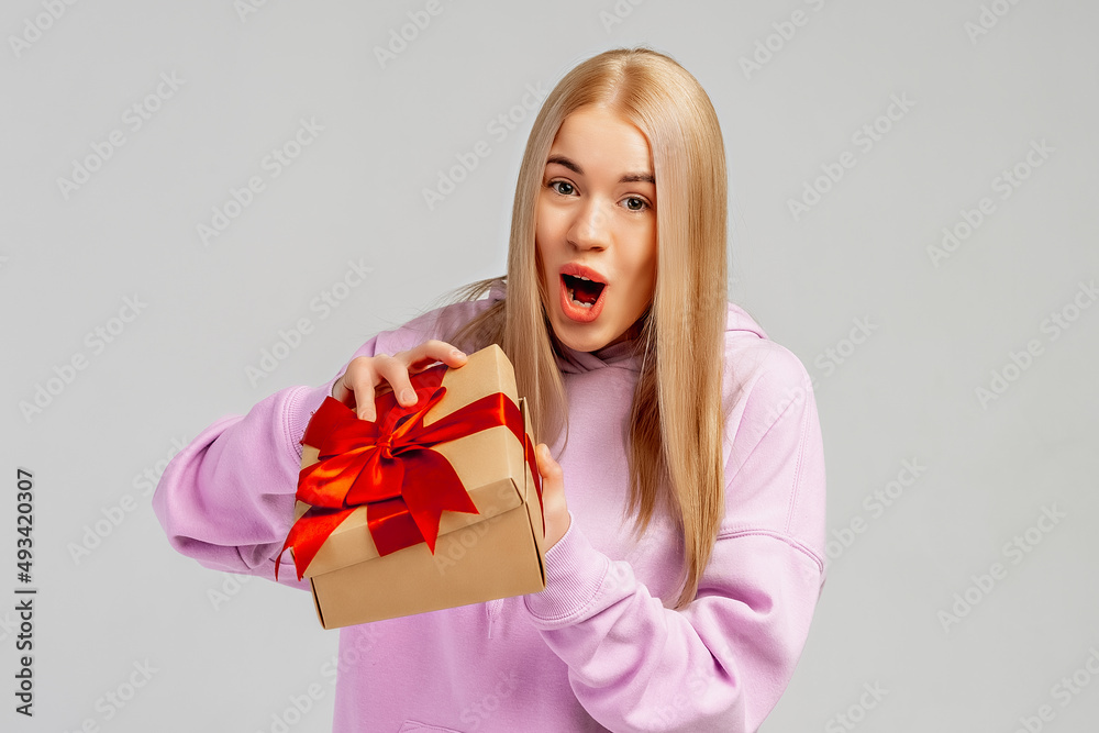 Excited young woman in trendy hoodie opening present gift box. Pretty girl smiling and posing with gift box on light background. New Year Women's Day birthday holiday concept Excited young woman in trendy hoodie opening present gift box. Pretty girl smiling and posing with gift box on light background. New Year Women's Day birthday holiday concept