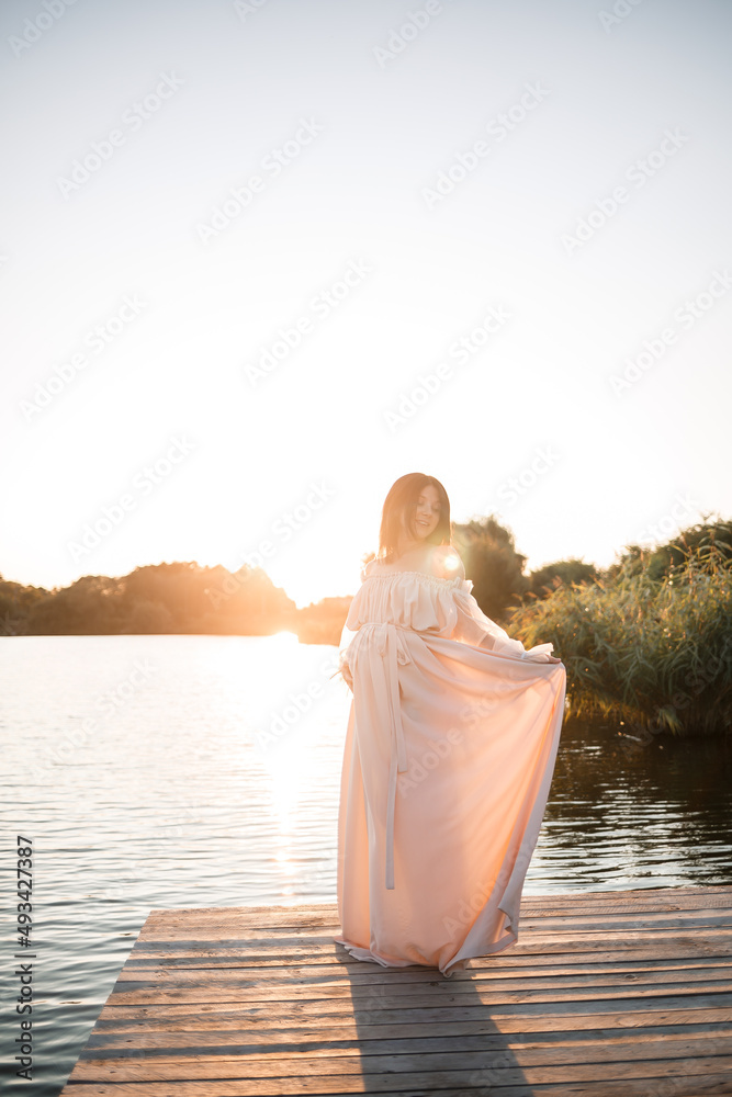 A young pregnant woman in a chiffon dress stands on a pier by the river against the backdrop of an orange sunset