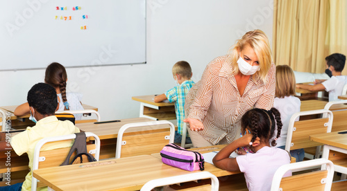 Young female teacher in protective face mask giving lesson to children in elementary school. New life reality during coronavirus pandemic © JackF