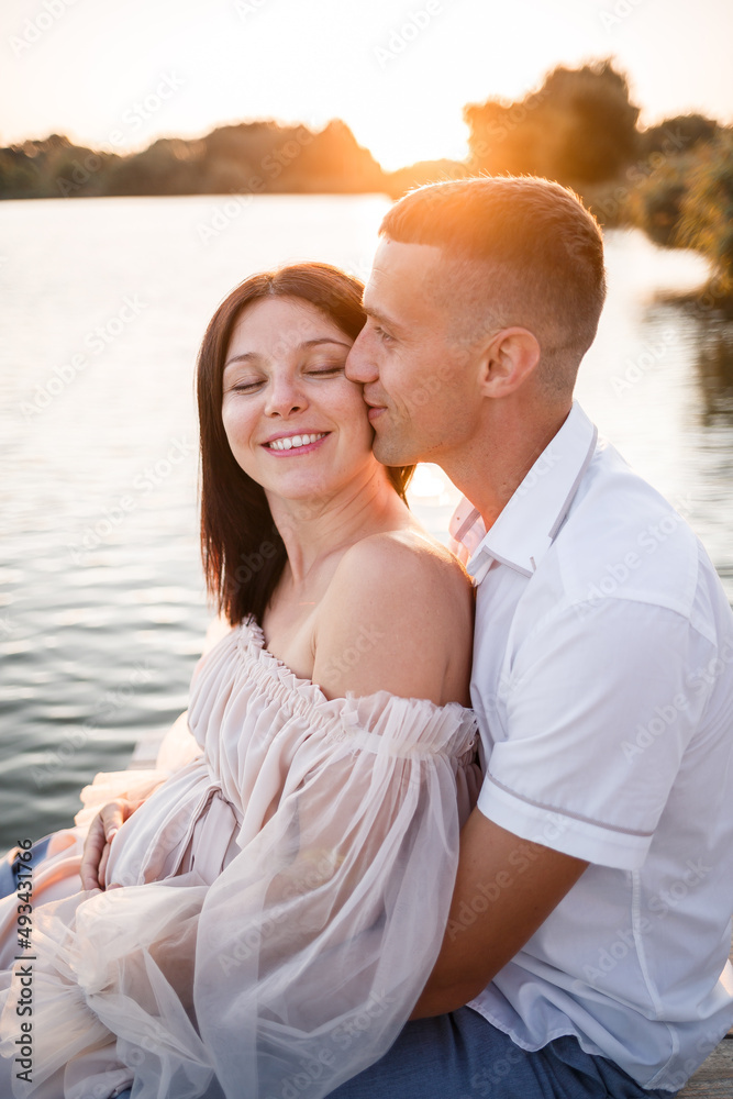 A husband with a pregnant wife is resting on the river bank, sitting on a pier in summer sunny weather at sunset. couple in love man and woman hugging, holding hands, A husband with a pregnant wife is resting on the river bank, sitting on a pier in summer sunny weather at sunset. couple in love man and woman hugging, holding hands,