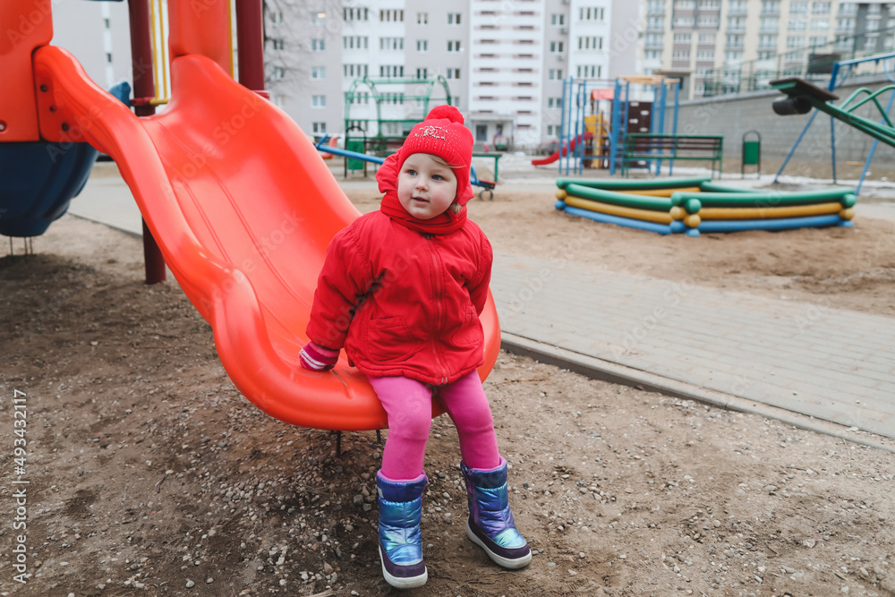 Happy girl in a red jacket rides on a hill at the playground. funny cheerful toddler girl playing outdoors