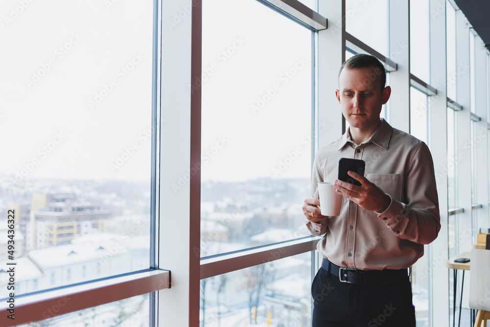 Male brokers with phone looking towards window with coffee in hand in modern workspace Male brokers with phone looking towards window with coffee in hand in modern workspace
