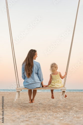 Young mother and little fair-haired girl sit on swing on sea background during sunset. Mom and daughter on the beach over yellow magic sunset lights.